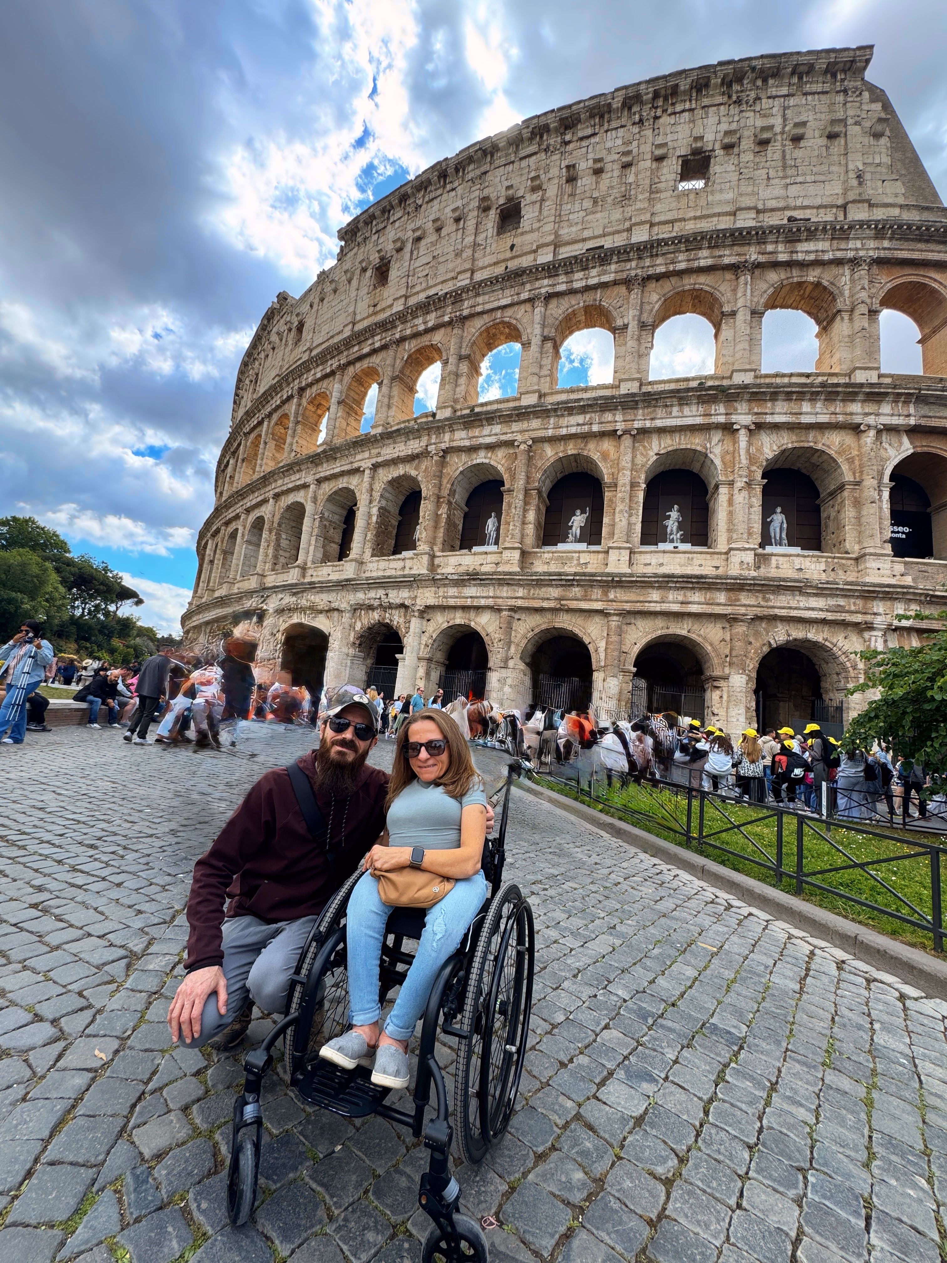 Heather is in her wheelchair and is next to her husband Dustin, who both pose together on the cobblestone plaza in front of the Colosseum in Rome. Heather is wearing sunglasses, a light blue tshirt, and jeans, while Dustin wears a burgundy sweatshirt and sunglasses. The ancient Roman amphitheater rises dramatically behind them against a partly cloudy blue sky, with tourists visible around the monument's base.